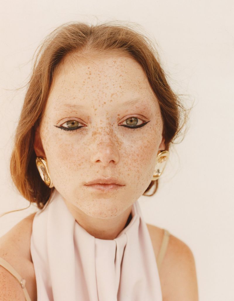 Close-up portrait of model Sara Grace Wallerstedt with freckles, winged black eyeliner, and gold earrings wearing a pale top.