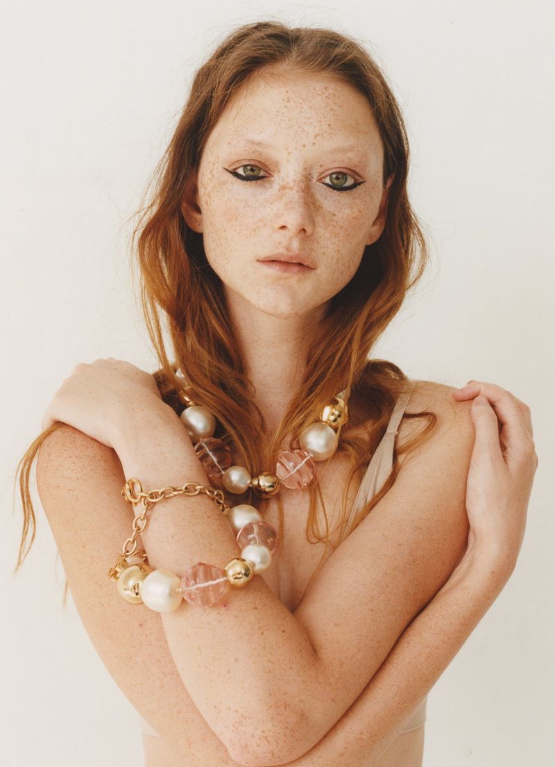 Portrait of a red-haired young adult woman with freckles wearing a pearl and gold necklace and bracelet against a white backdrop.