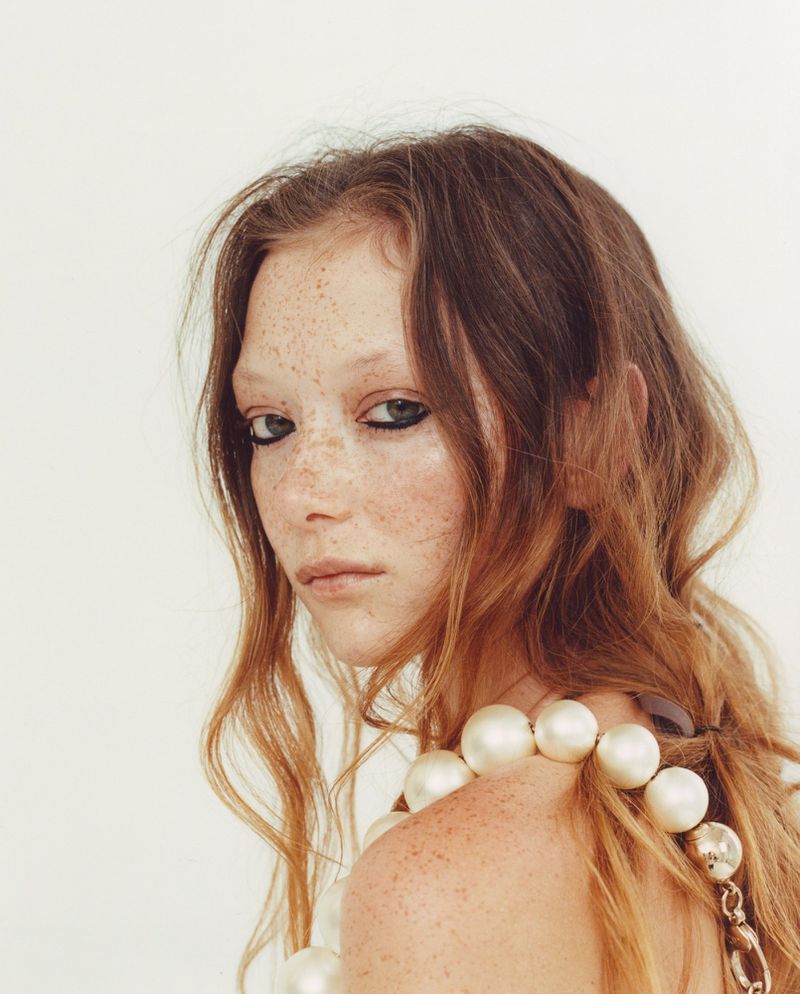 Close-up of a woman with freckles and auburn hair wearing a large pearl necklace in a beauty and fashion shot against a white backdrop.