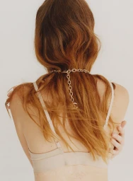 Model seen from behind with long auburn hair wearing a beige bra and a chunky gold chain necklace against a light background