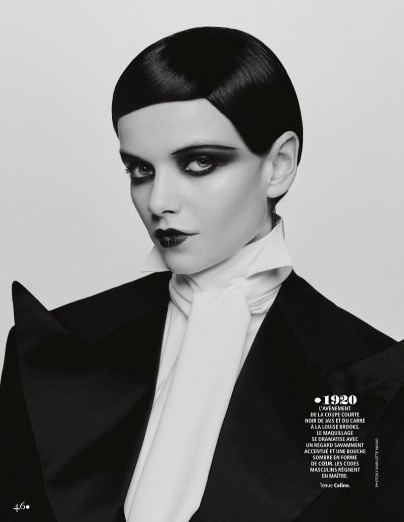 Monochrome portrait of a woman with dark, short hair, heavy eye makeup, and dark lipstick, wearing a black blazer and white ascot indoors.