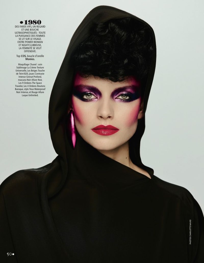 Portrait of a woman with bold pink and purple makeup wearing a hooded black garment against a plain background in a studio shot.