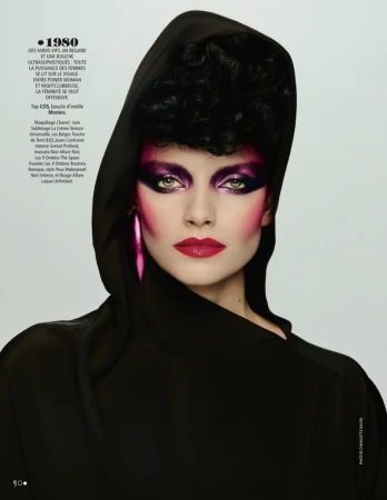 Portrait of a woman with bold pink and purple makeup wearing a hooded black garment against a plain background in a studio shot.
