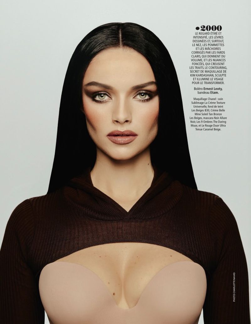 Close-up studio shot of a brunette woman with smokey eye makeup wearing a brown cropped hooded sweater and a beige strapless top.