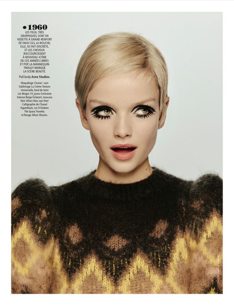 Close-up fashion shot: young adult woman with short blonde hair and heavy black eye makeup, wearing a black and brown patterned sweater.