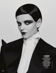 Black and white portrait of a woman with slicked hair, dramatic dark eye makeup, and a black suit jacket with white necktie.