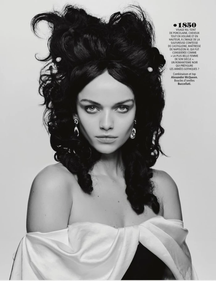 Model with voluminous dark historic hairstyle and pearl hairpins wearing an off-shoulder white top against a grey backdrop
