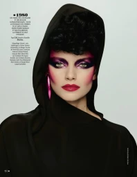 Model with bold purple eyeshadow and pink blush wearing a black hooded top against a grey seamless background.
