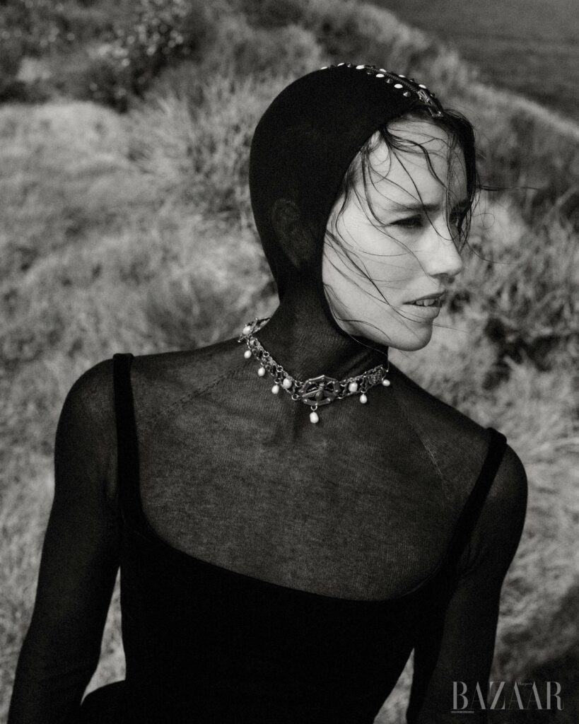 Monochrome portrait of a woman with a net head covering and pearl necklace, set against a grassy hillside backdrop, high fashion look.