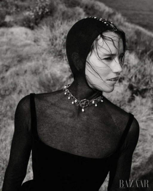Monochrome portrait of a woman with a net head covering and pearl necklace, set against a grassy hillside backdrop, high fashion look.