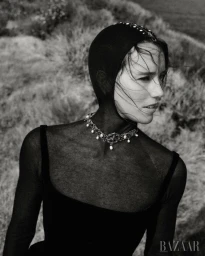 Black and white portrait of a woman wearing a dark fitted hood and ornate pearl necklace against an outdoor landscape background.