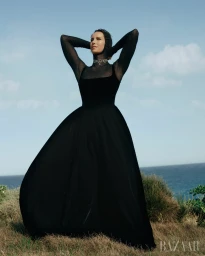 Woman wearing a black long-sleeved dress and head covering standing outdoors on a grassy coastal hill against an ocean horizon.