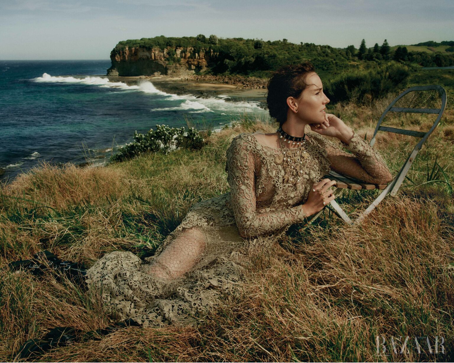 A woman in a long lace gown is lying in a grassy field near a rocky coastline on a sunny day, with a chair nearby and the ocean in the distance.