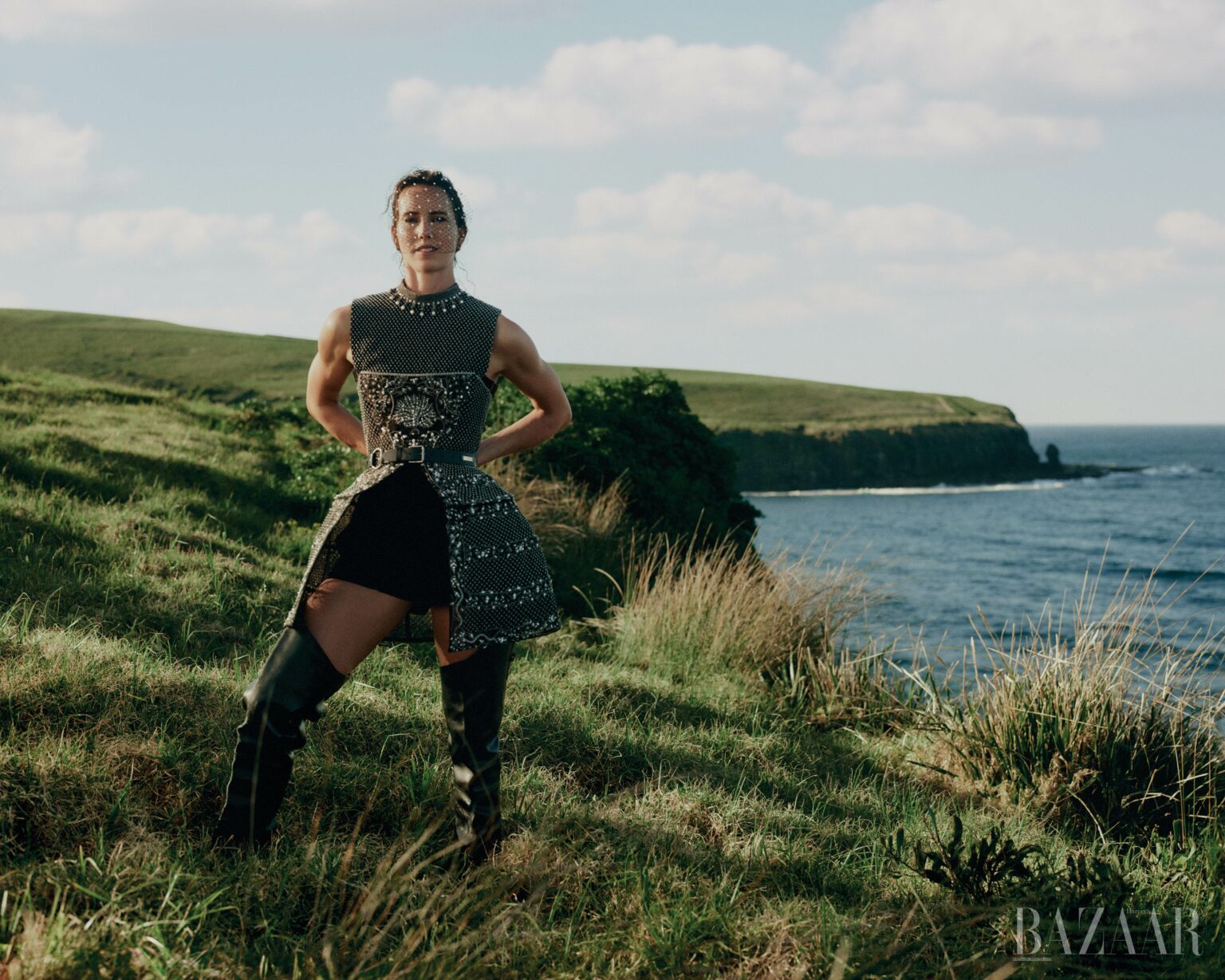 Woman standing on a grassy coastal cliff overlooking the sea, wearing a patterned sleeveless top and tall dark boots.