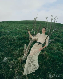 Woman in an ornate lace dress leaning against a bare tree on a grassy hillside under a pale sky