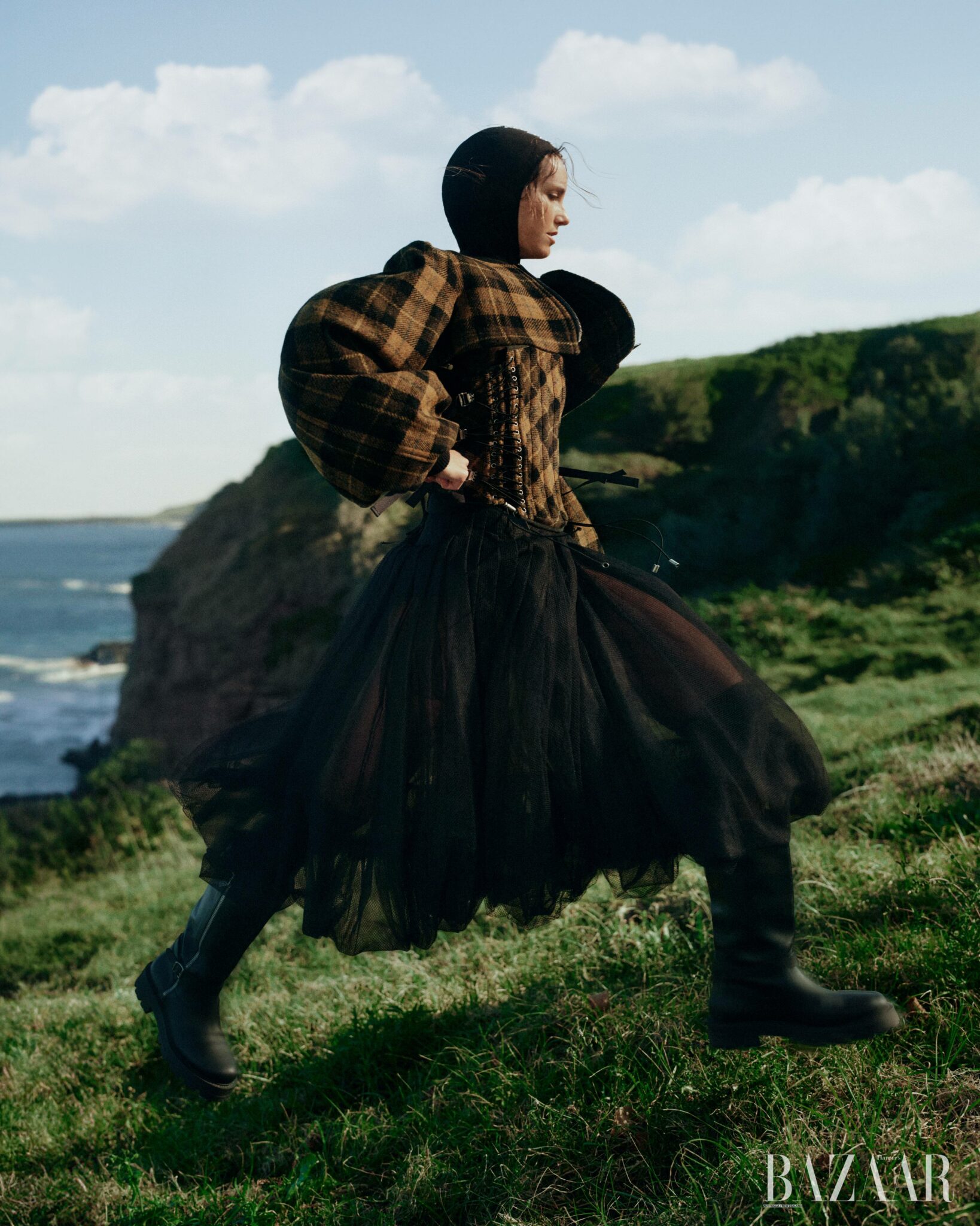 Woman in plaid puffed-sleeve top and black tulle skirt running along a coastal grassy cliffside
