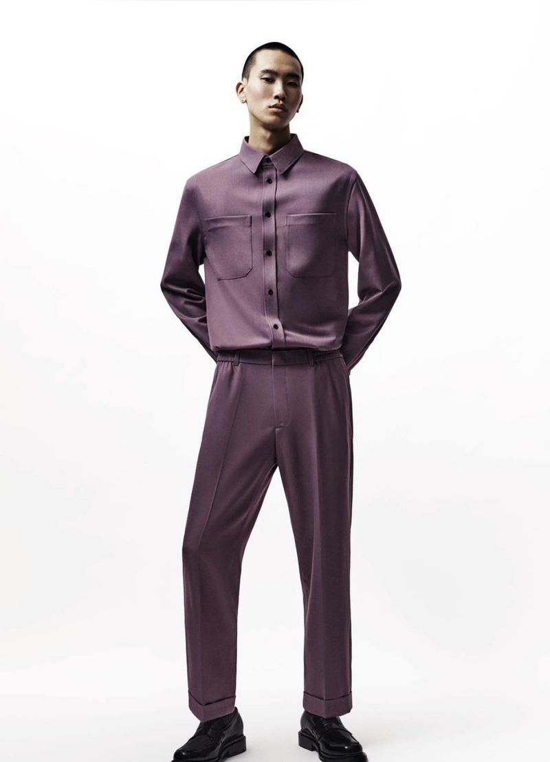 A stylish male model poses in a light purple, long-sleeved button-down shirt and matching trousers against a stark white background.
