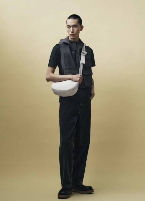 Full-length shot of an adult Asian male wearing a black t-shirt, dark vest, pants, and shoes, carrying a white crossbody bag against a pale yellow background.