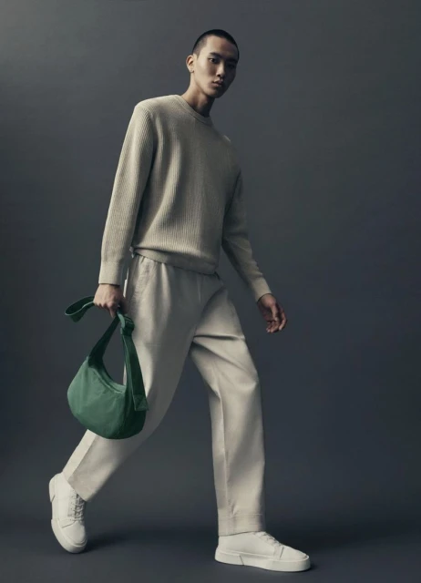An Asian male model walks in a studio wearing a beige knit sweater, matching pants, white sneakers, and carrying a green bag.
