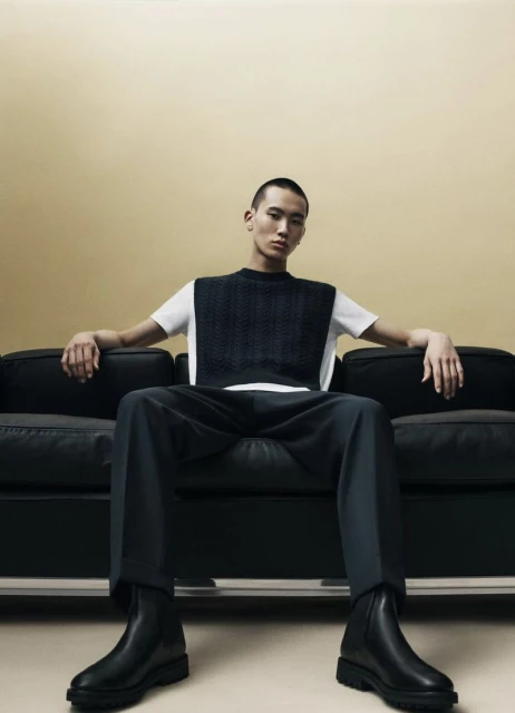 An adult male sits on a black couch wearing a white t-shirt, dark grey sweater vest, matching pants, and black boots, against a cream background.