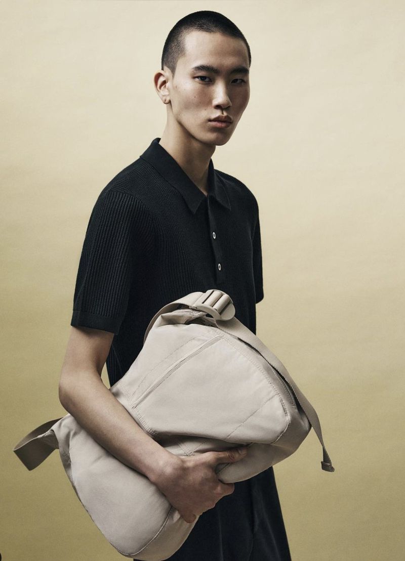 Young Asian male fashion model with a shaved head poses with a large beige shoulder bag against a neutral yellow background.