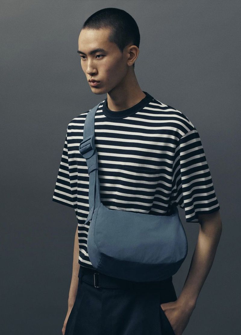 Studio shot of a young adult man wearing a striped t-shirt and a blue crossbody bag against a grey background.