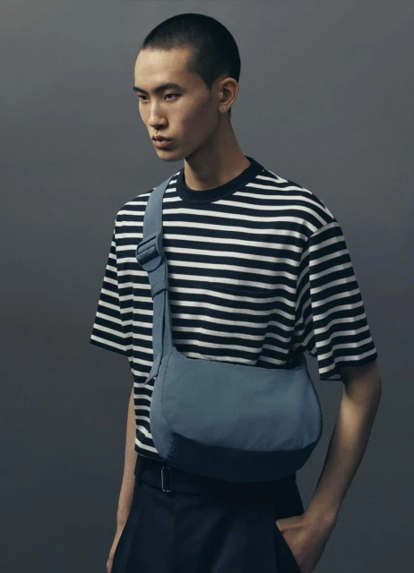 Studio shot of a young adult man wearing a striped t-shirt and a blue crossbody bag against a grey background.
