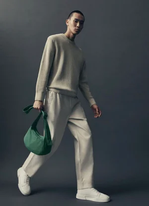 Model wearing neutral knit sweater and trousers holding a green handbag against a dark studio backdrop