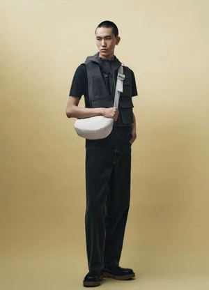 Model wearing dark utility vest and trousers holding a light crossbody bag against a neutral studio backdrop