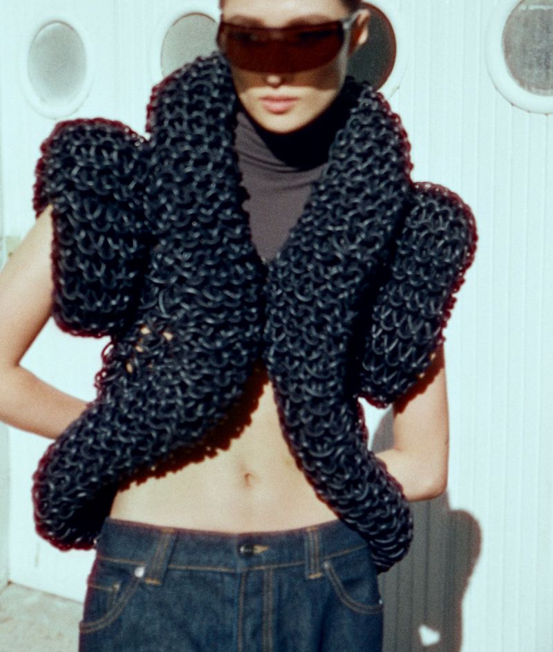 Woman poses in a futuristic oversized black knit vest, wearing tinted sunglasses, dark turtleneck, and dark wash denim jeans against a white wall.