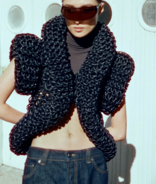 Woman poses in a futuristic oversized black knit vest, wearing tinted sunglasses, dark turtleneck, and dark wash denim jeans against a white wall.