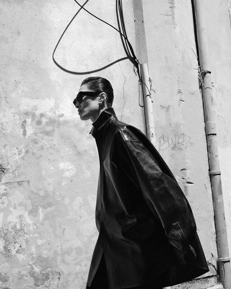 Black and white fashion shot of a female model in a black leather trench coat, sunglasses, posing against an urban wall backdrop.