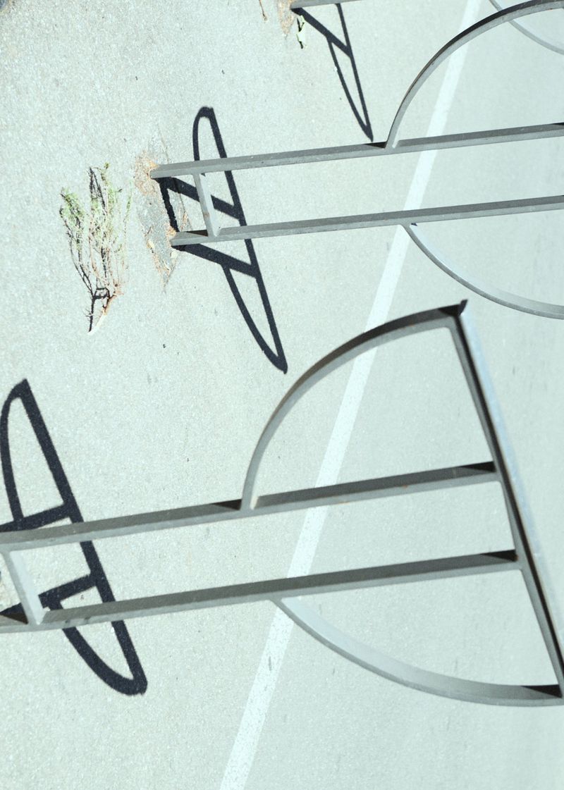 Abstract view of bike rack on concrete with shadows and partial weeds; horizontal orientation featuring minimal color palette.