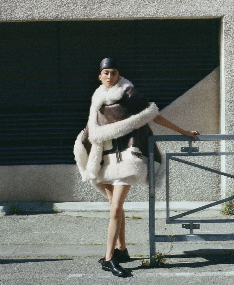 A woman in a stylish shearling-trimmed brown cape and black leather hat poses next to a metal barricade on a city street.