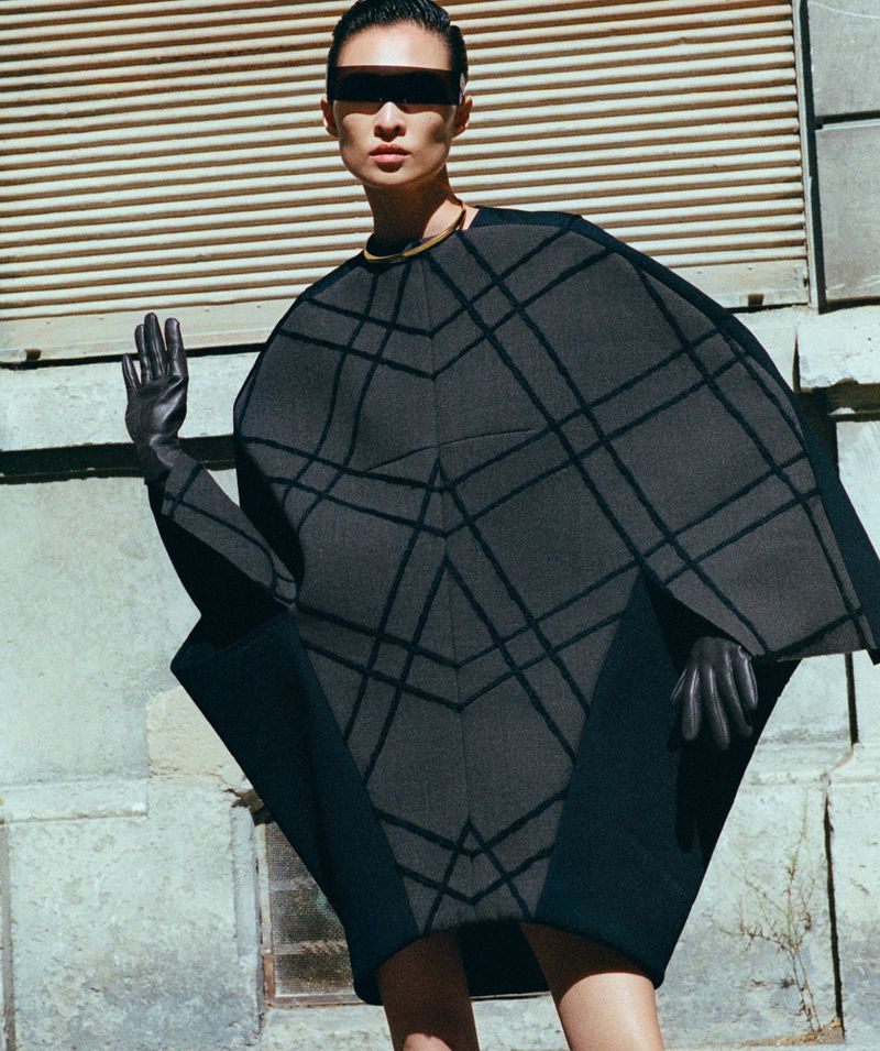 Model in architectural dark grey patterned poncho wearing dark sunglasses against an urban building wall