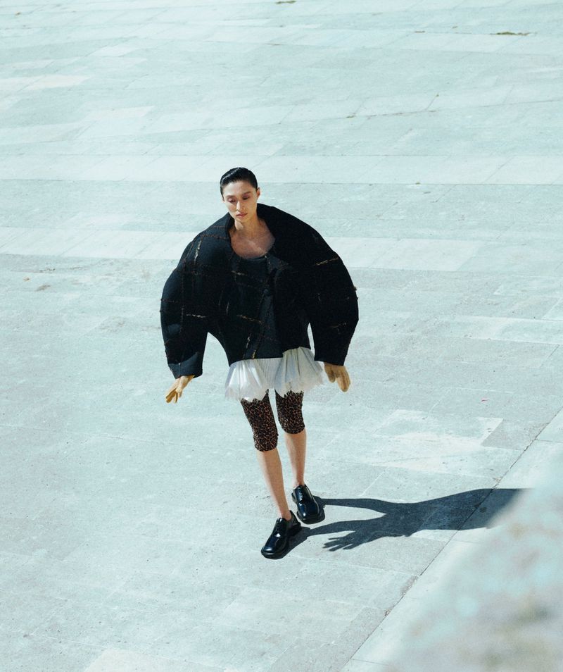 High-angle shot of a woman in an oversized black jacket, white frilly skirt, and printed leggings posing on a textured light-grey surface.