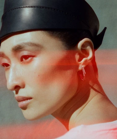 Close-up portrait of model Chu Wong wearing a black leather headpiece and hoop earring with a horizontal red light flare across her face.