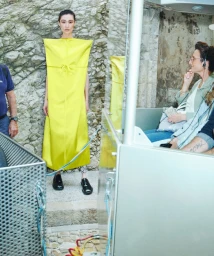 Model Chu Wong in a sculptural bright yellow rectangular dress standing beside a stone wall and a transport cabin.