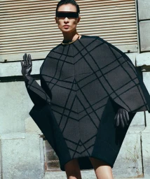 Model in architectural dark grey patterned poncho wearing dark sunglasses against an urban building wall
