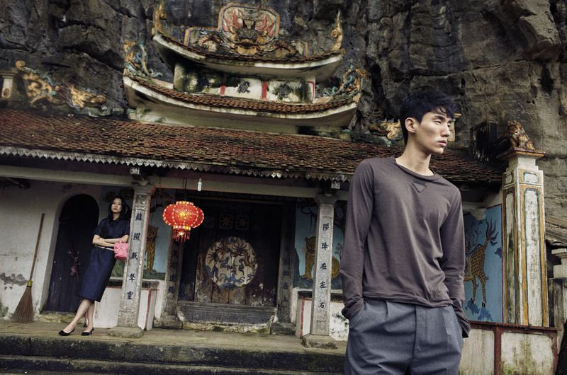 Asian male model poses in front of an old Asian temple structure with a woman in a navy dress and a red Chinese lantern.