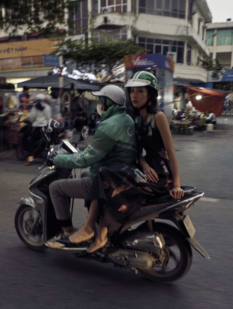 Two people ride a scooter through a busy city street. The woman wears a floral dress and green helmet, while the driver is in a green jacket.