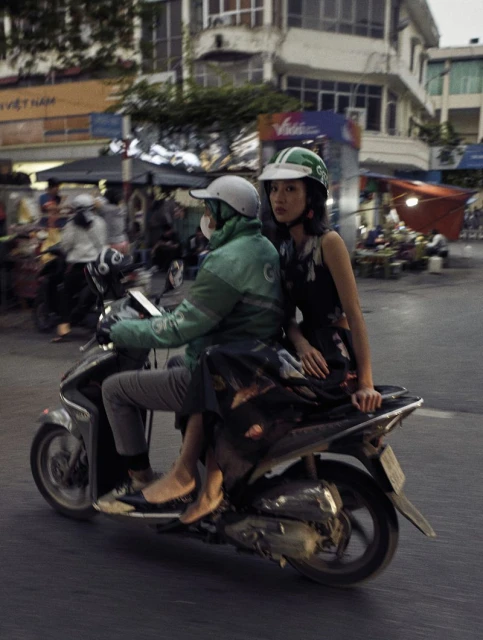 Two people ride a scooter through a busy city street. The woman wears a floral dress and green helmet, while the driver is in a green jacket.
