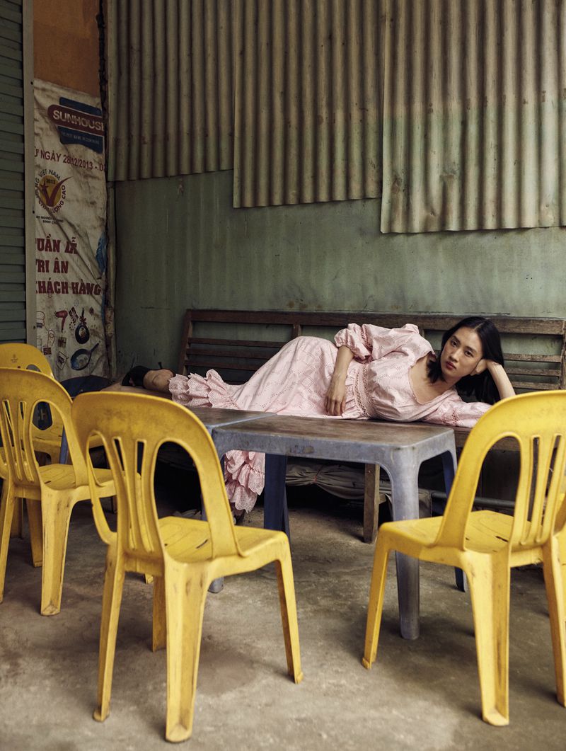Asian woman in a pink dress lying on a blue table surrounded by yellow chairs in an outdoor cafe with a textured metal wall backdrop.