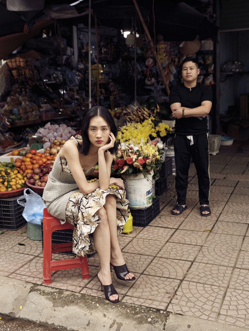 A woman poses on a red stool in a patterned dress, with a man standing in the background at a market filled with produce and flowers.