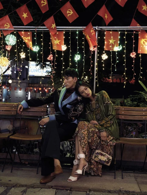 A stylish couple is sitting outside at night under string lights and red star flags. The woman leans on the man, who is looking away.