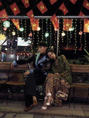 A stylish couple is sitting outside at night under string lights and red star flags. The woman leans on the man, who is looking away.
