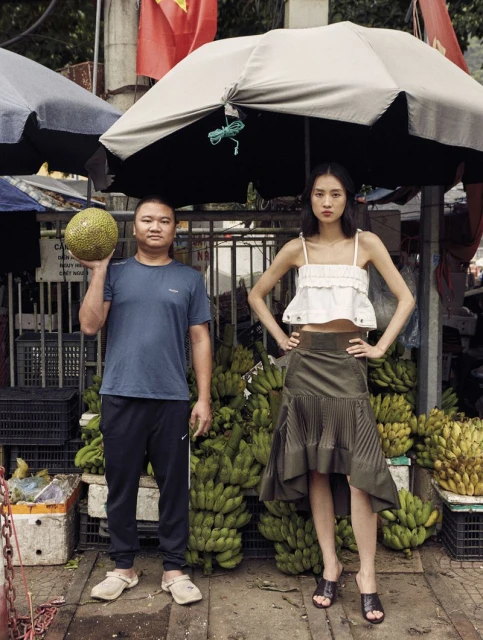 An Asian man holds jackfruit next to an Asian woman in a skirt and top standing in front of bananas in an outdoor market.