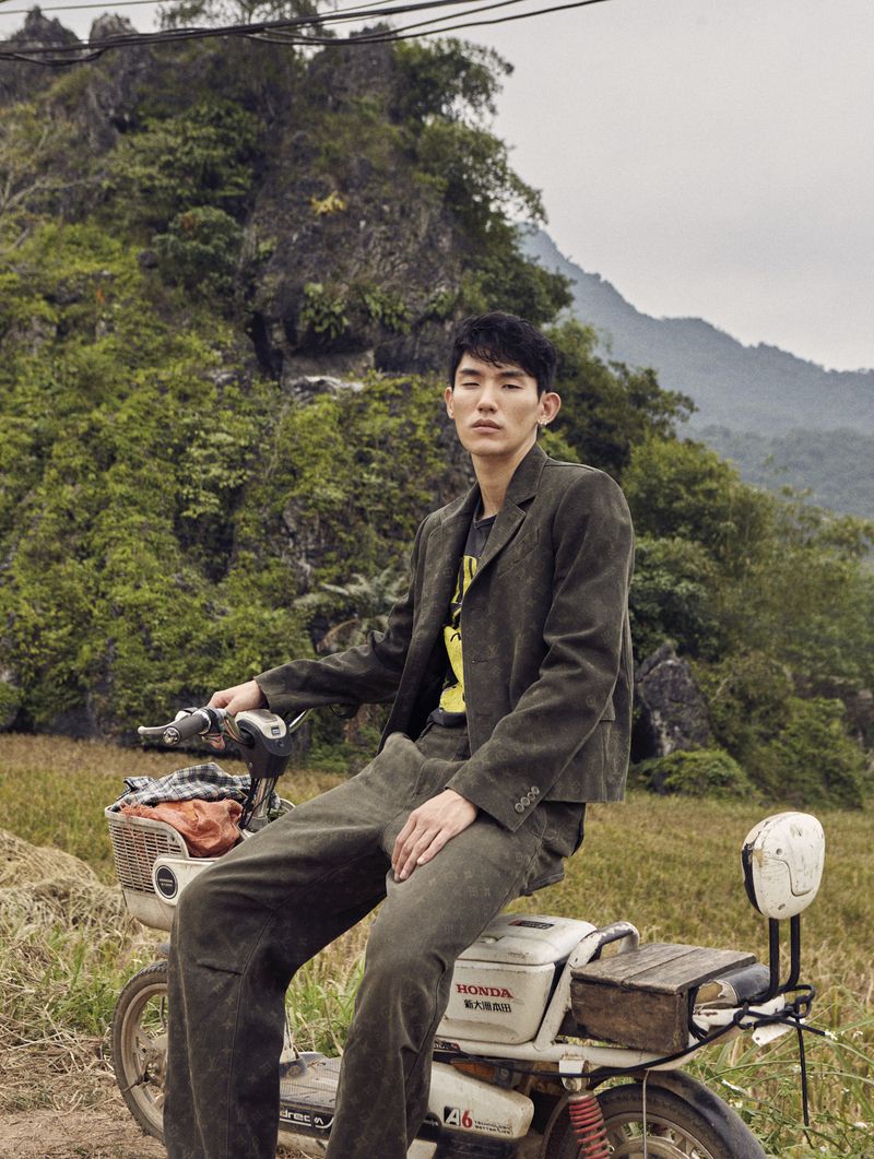 Asian male model sitting on an old moped, wearing a corduroy suit in a rural landscape with lush green mountains in the background.