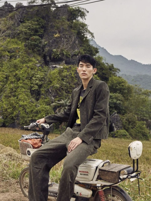 Asian male model sitting on an old moped, wearing a corduroy suit in a rural landscape with lush green mountains in the background.