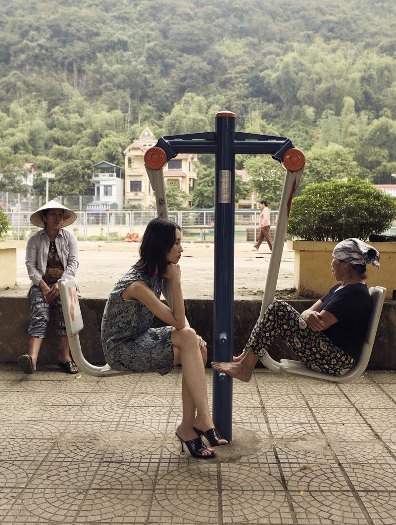 Three women seated on outdoor fitness equipment in a park setting with a lush green mountain backdrop and nearby buildings.